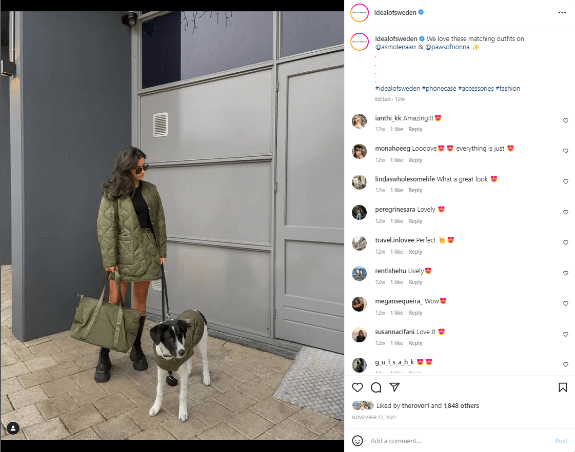 A woman and her dog stand outside a gray building in matching green jackets. The woman holds a green, oversized purse. 